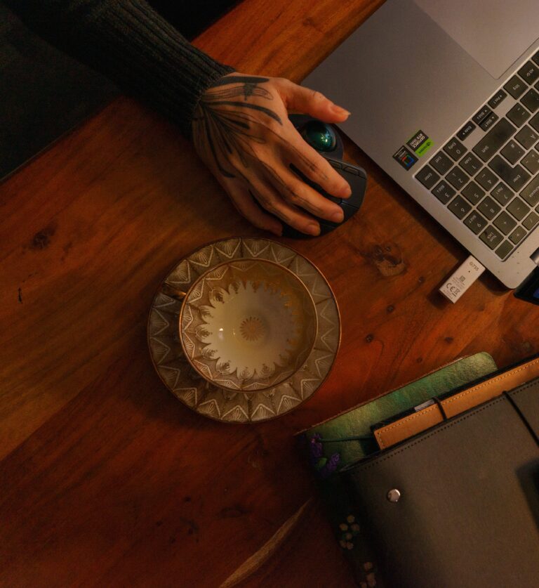 Eine goldene Sammeltasse vor einem Stapel Notizbücher. Im Hintergrund ist ein Laptop zu sehen. Eine stark tätowierte Hand hält die Maus des Laptops.